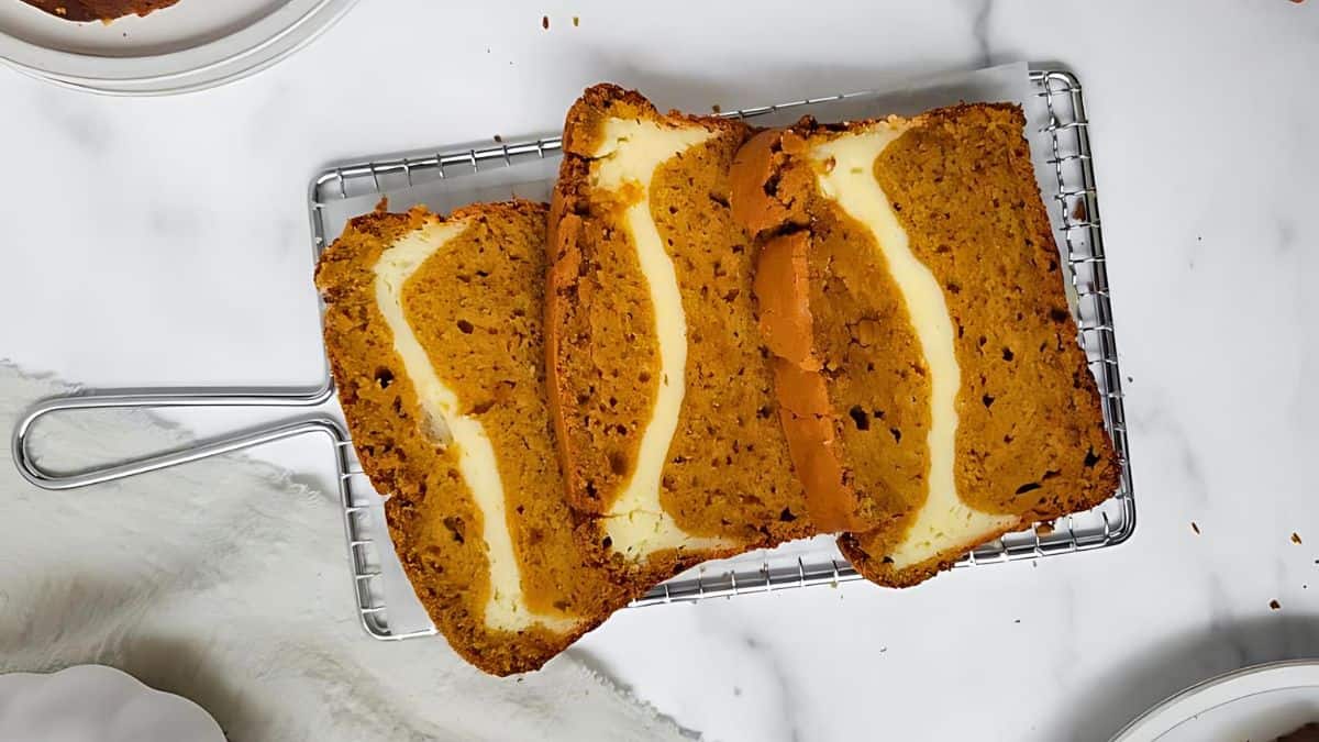 Three slices of pumpkin bread with a cream cheese filling are arranged on a wire cooling rack over a white surface. The bread has a golden-brown crust and a rich, moist texture, with swirls of creamy filling clearly visible in each slice.