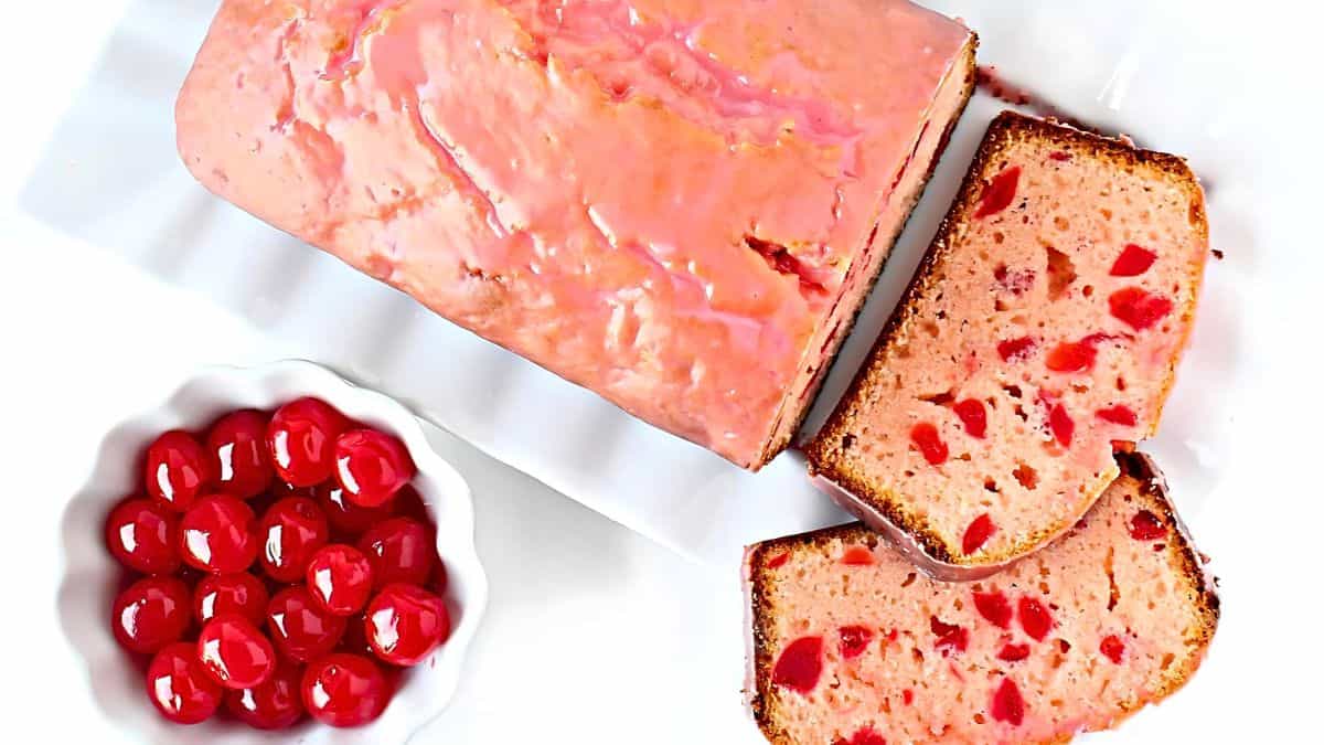 A sliced glazed cherry loaf cake is displayed on a white rectangular plate. Beside the plate is a white bowl filled with bright red maraschino cherries. The cake has chunks of cherries visible in the slices.