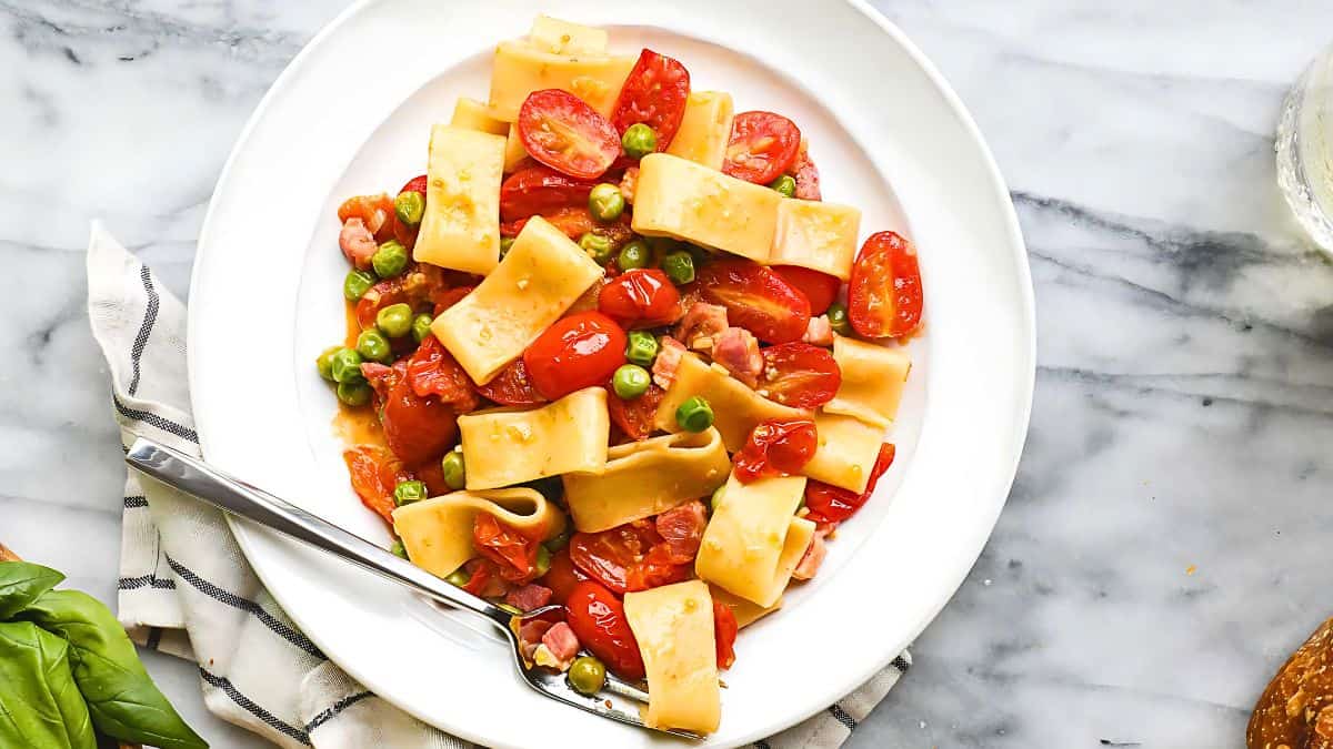 A white plate filled with paccheri pasta, cherry tomatoes, green peas, and diced ham. The dish is garnished with grated cheese. A fork rests on the left side of the plate, and a striped napkin is partially visible underneath. The background is a marble surface.