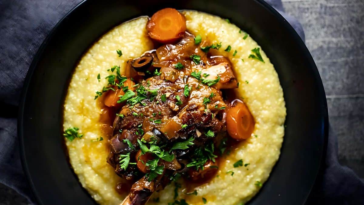 A bowl of creamy polenta topped with a braised lamb shank, garnished with chopped parsley, cooked carrots, and a rich sauce. The dish is presented on a dark plate with a rustic background.