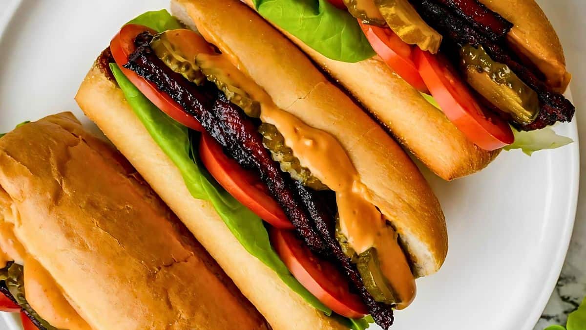 Three freshly made vegan BLT sandwiches on toasted buns are displayed on a white plate. The sandwiches are filled with crispy tempeh bacon strips, lettuce, tomato slices, pickles, and a generous spread of creamy sauce. The vibrant colors of the ingredients are visible.