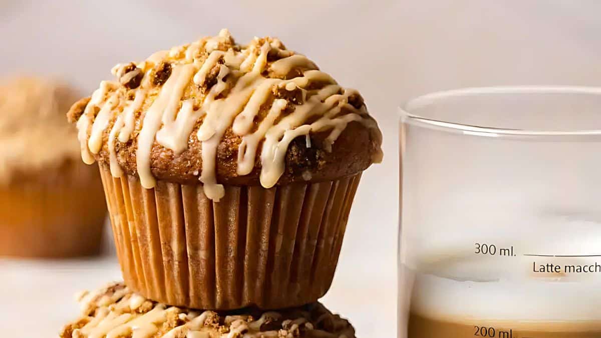 Close-up of a muffin drizzled with icing, sitting on top of another muffin. In the background, there is a glass filled with a creamy beverage marked with measurement lines. Another muffin is slightly blurred in the distance, giving a cozy and inviting feel.