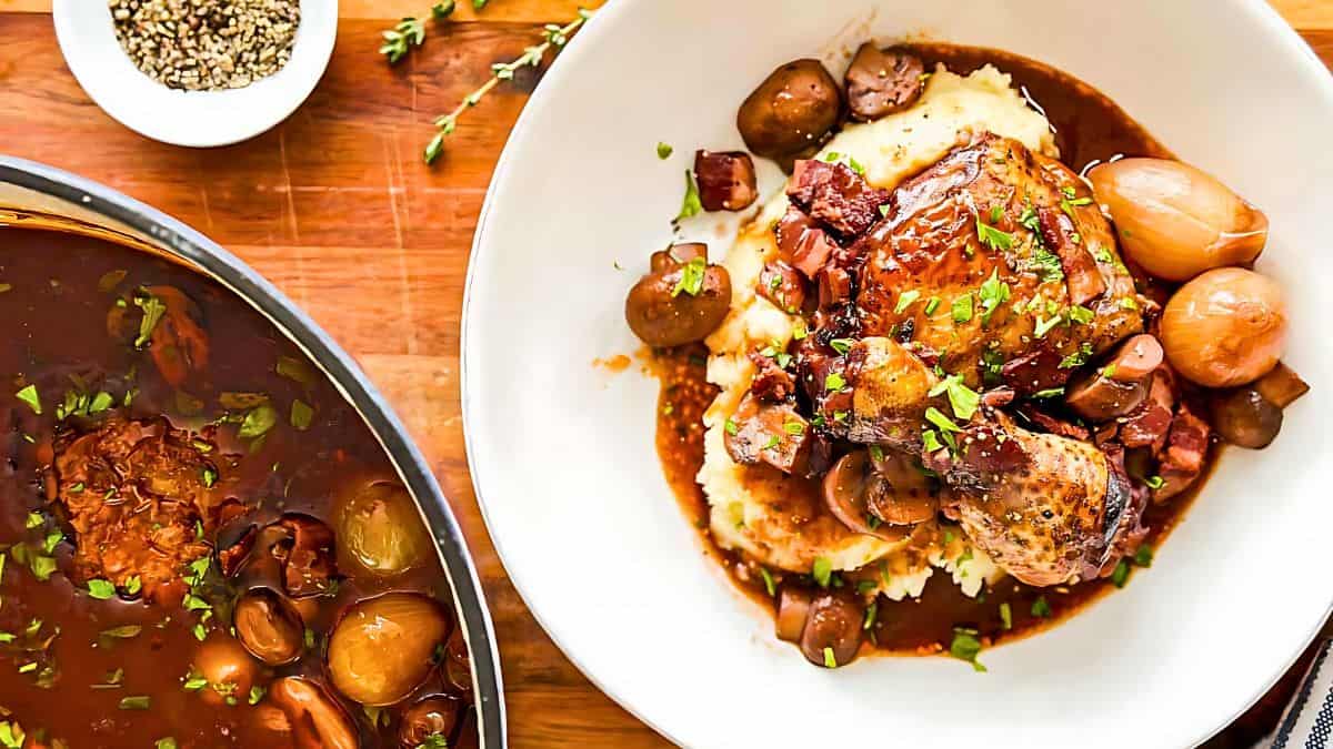 A white bowl filled with coq au vin sits on a wooden surface. The dish features chicken, mushrooms, pearl onions, and a rich wine sauce over mashed potatoes. Fresh herbs garnish the meal. Nearby, a pot of the same dish and a small bowl of pepper are visible.