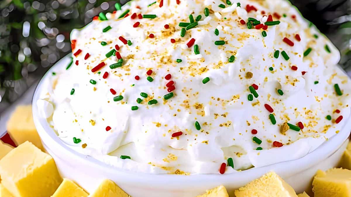 A close-up of a festive bowl of white dip topped with red and green sprinkles, surrounded by small, cubed pieces of yellow bread. The dip appears fluffy with a hint of gold-colored seasoning sprinkled on top. The background is blurred with hints of greenery.