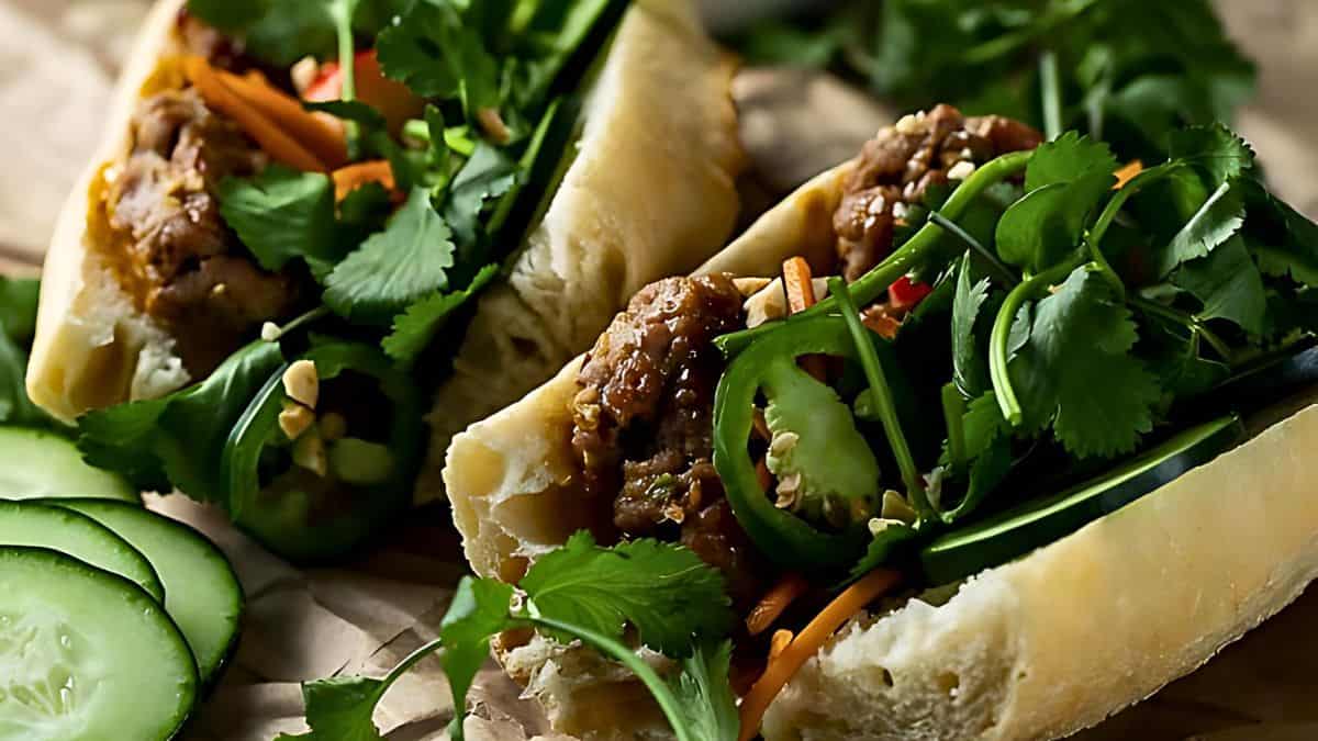 A close-up view of two fresh bánh mì sandwiches filled with grilled meat, cilantro, julienned carrots, sliced cucumbers, green onions, and jalapeño peppers. The sandwiches are placed on parchment paper with extra cucumber slices nearby.