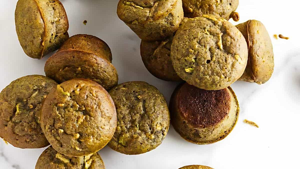 A pile of golden-brown muffins with slightly cracked tops, showing flecks of darker ingredients, is scattered across a white marble surface. Some muffins are lying on their sides, showcasing their rounded edges and moist texture.