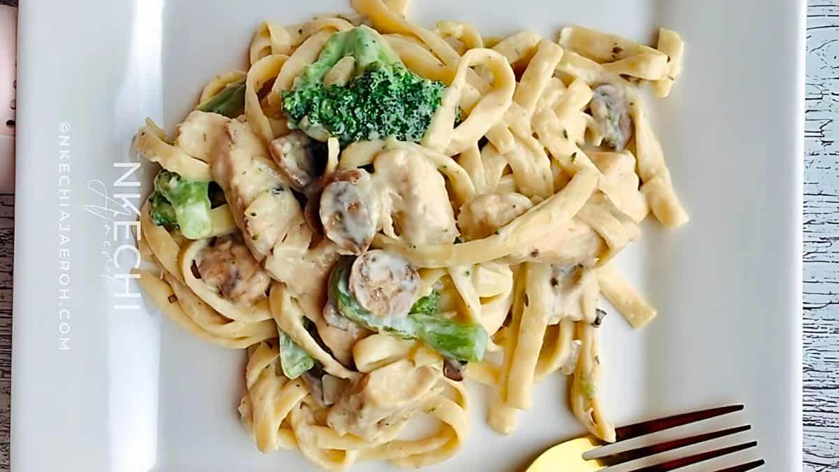 A square white plate holds a serving of creamy fettuccine alfredo mixed with broccoli and mushrooms. The dish appears garnished with herbs. A gold fork is placed on the right side of the plate.