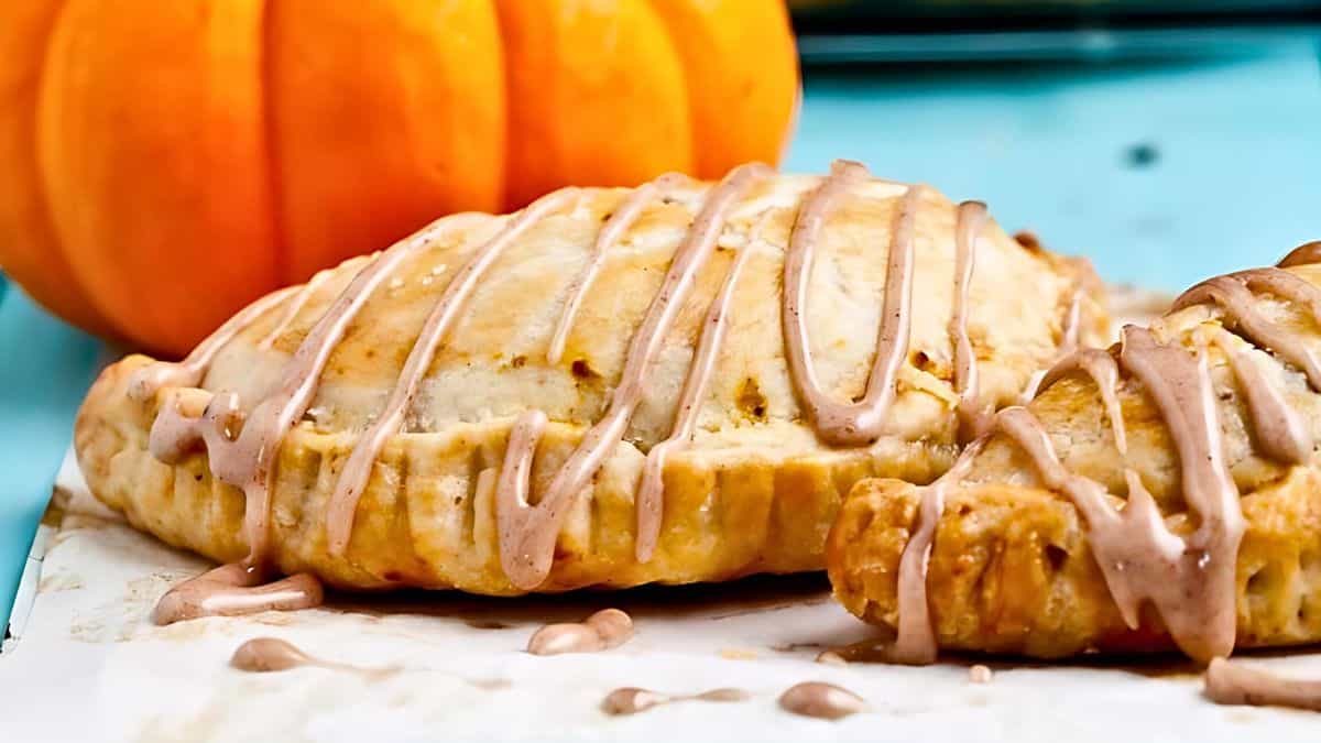 Close-up of two pumpkin hand pies drizzled with a light glaze on parchment paper. The background features a small pumpkin, adding an autumnal touch. The pastries have a golden-brown crust with the glaze artistically swirled on top.