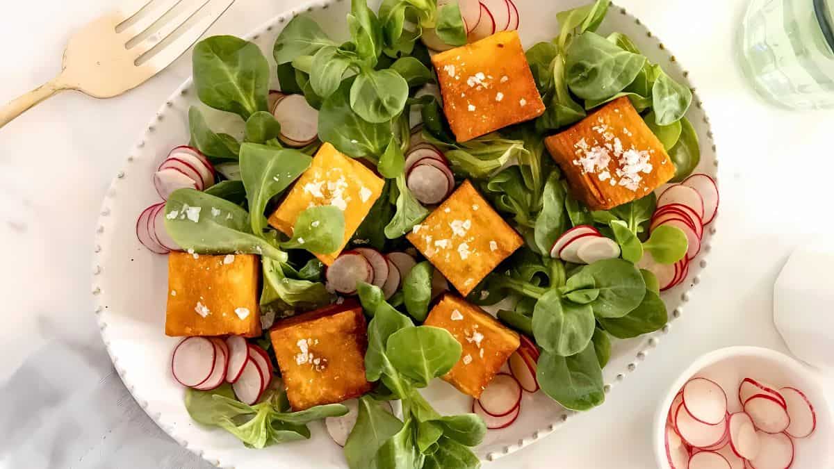 A fresh salad on an oval white plate features cubed roasted butternut squash on a bed of leafy greens, garnished with sliced radishes and salt flakes. A fork is visible above the plate, and a glass of water is partially seen to the right.