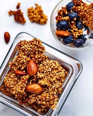 A glass jar brimming with crunchy granola clusters and almonds sits on a white surface. Nearby, a bowl with granola, blueberries, and a spoon awaits—a perfect make-ahead breakfast to kickstart your day with wholesome goodness.