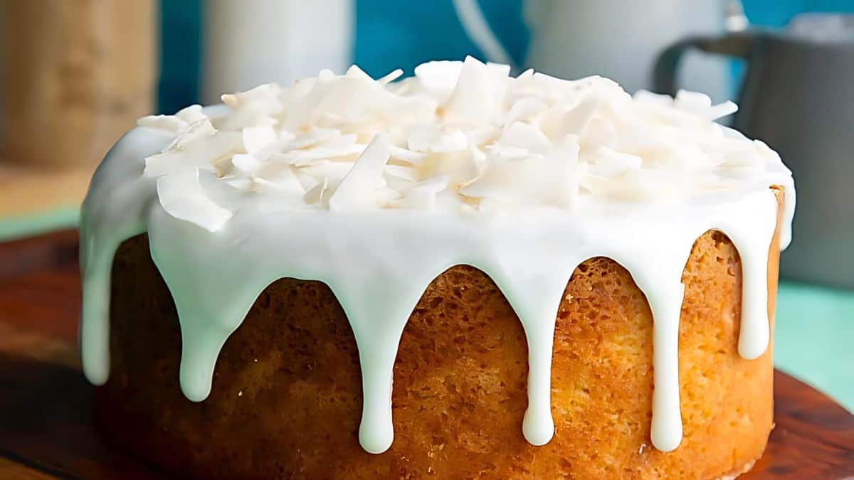 A round, golden-brown cake topped with dripping white icing and garnished with large, shaved coconut pieces sits on a wooden surface. In the background, there's a blurred blue and teal backdrop with some containers visible.