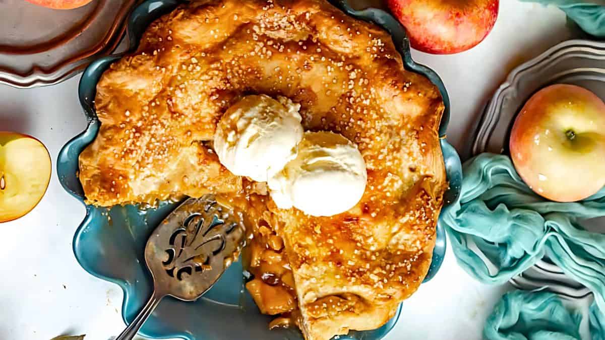 A partially sliced golden-brown pie in a blue dish, topped with two scoops of vanilla ice cream. A silver pie server rests nearby. Surrounding the pie are whole apples and a light blue cloth, creating a cozy and inviting setting.