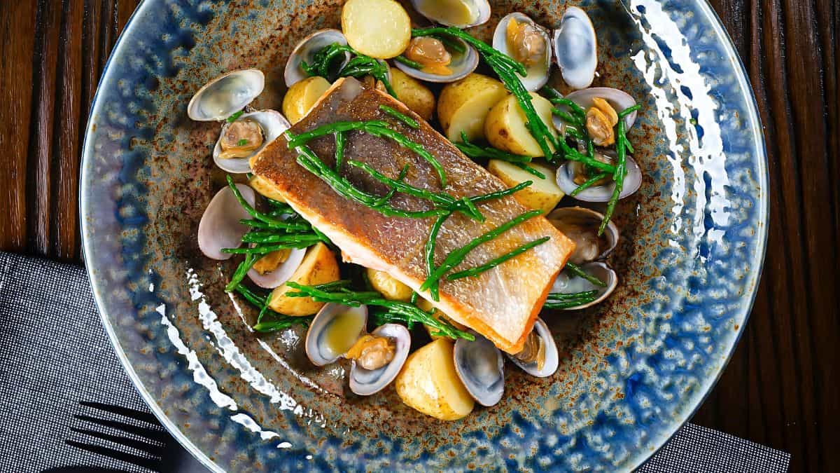 A beautifully plated dish featuring a piece of seared fish garnished with green herbs, served alongside baby potatoes and clams on a speckled blue ceramic plate. The food is arranged artfully with a textured wooden surface in the background.