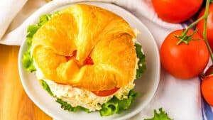 A croissant sandwich filled with chicken salad, lettuce, and tomato is placed on a white plate. Some fresh tomatoes are arranged next to it, along with a white napkin partially visible in the background.