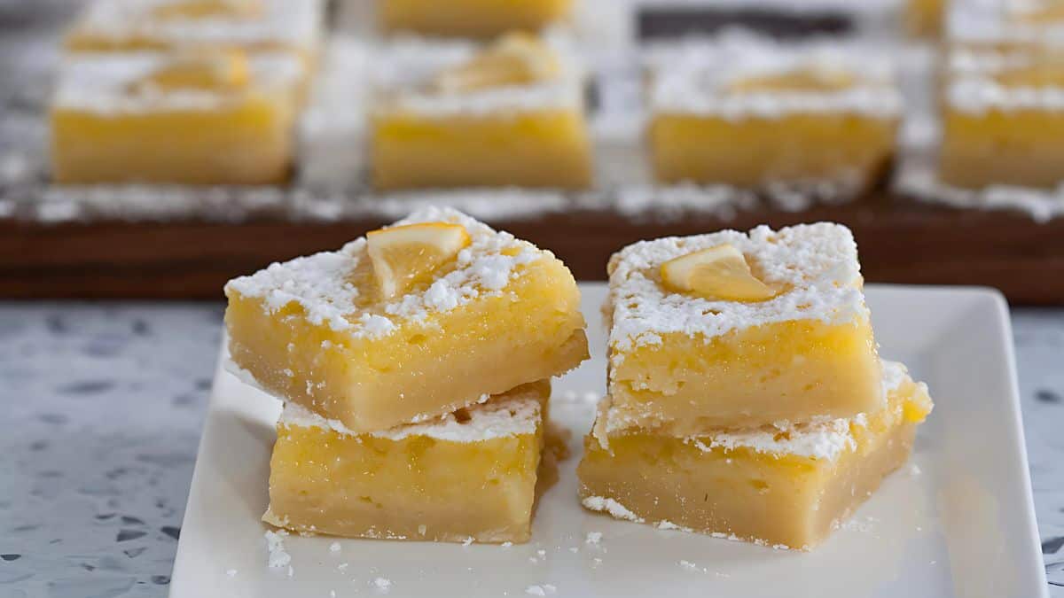 Three lemon bars are stacked on a white plate, dusted with powdered sugar and topped with small lemon wedges. More lemon bars are visible in the background on a wooden board. The surface beneath the plate is speckled.
