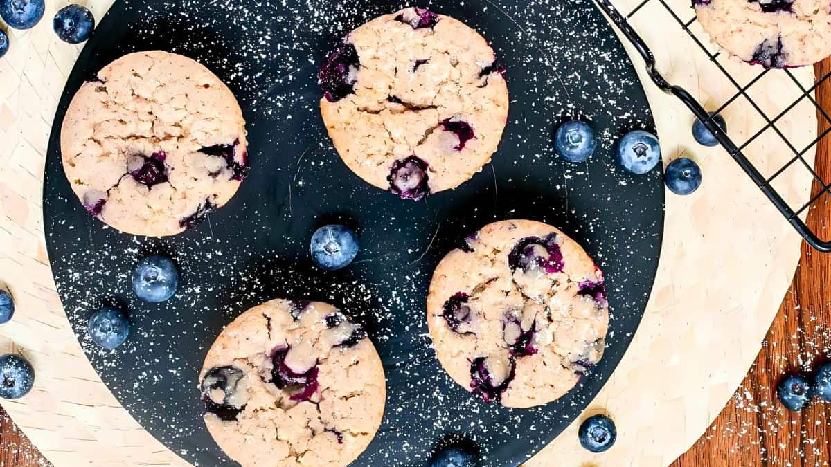Four blueberry muffins rest on a round, dark board. Fresh blueberries are scattered around the muffins, some on the board and others on a wooden surface nearby. A cooling rack with another muffin and more blueberries is partially visible on the right.
