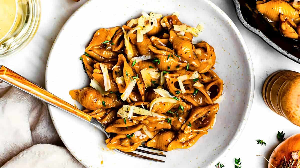 A white plate filled with creamy, saucy shell pasta, garnished with grated cheese and herbs. A fork rests on the plate. A pepper grinder, a partially visible pan of pasta, and a glass of white wine appear beside the plate.