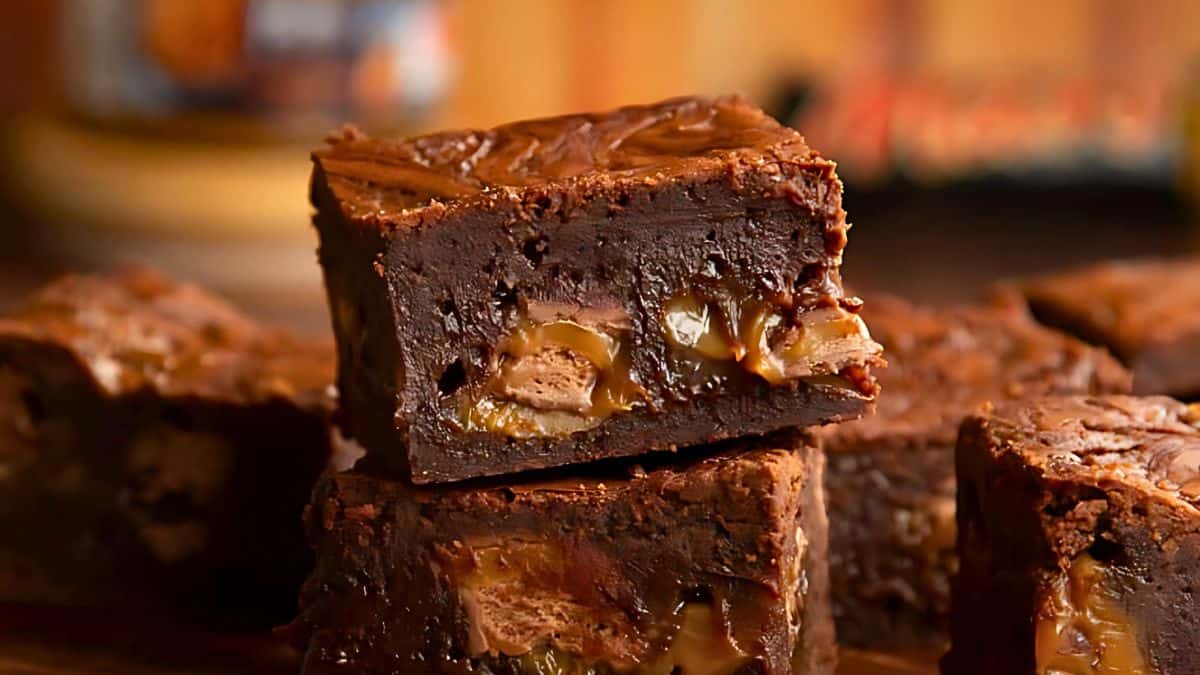 Close-up of decadent chocolate brownies stacked on top of each other. The brownies are rich and gooey, featuring a swirl of caramel and pieces of chocolate seamlessly blended into the dark fudge layers. They appear moist and full of texture, appealing to the sweet tooth.