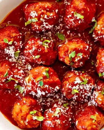 A bowl of tantalizing meatballs in rich tomato sauce, garnished with grated cheese and fresh herbs, makes for delightful meatball recipes for appetizers. The dish sits elegantly on a white tabletop, accompanied by a sprig of green basil on the side.