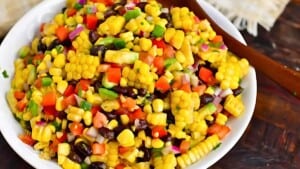 A vibrant corn salad featuring yellow corn kernels, black beans, chopped red bell peppers, red onions, diced cucumbers, and cilantro, all mixed in a white bowl. A wooden spoon rests on the edge of the bowl. The background shows a dark wooden surface.