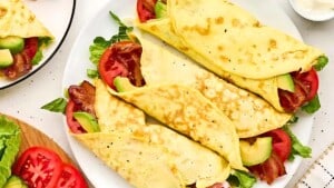 A plate of neatly folded crepes filled with crispy bacon, fresh avocado slices, and vibrant tomato slices, garnished with lettuce leaves. Surrounding the plate are additional toppings and ingredients on a white countertop.