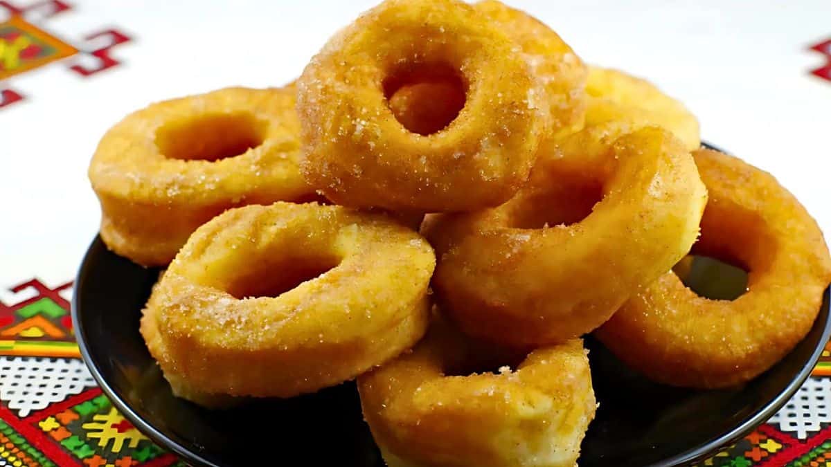 A plate of golden, sugar-coated donuts is stacked atop a colorful decorative fabric with a traditional pattern. The donuts appear freshly fried, with a slightly crisp outer layer and a soft, fluffy inside.