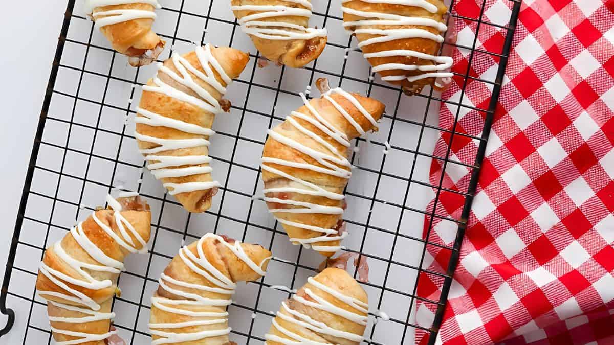 Freshly baked croissants drizzled with white icing are arranged on a cooling rack. The rack sits next to a red and white checkered cloth. The background is a white surface.
