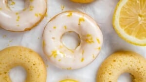 A close-up view of glazed lemon donuts placed on a white surface. Some donuts are glazed with a light lemon icing, garnished with lemon zest, while others are plain. A sliced lemon is visible in the upper right corner.