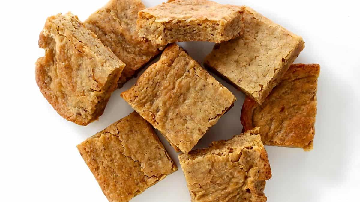 A close-up of several square-shaped blondie brownies arranged on a white surface. The blondies have a golden-brown, slightly cracked top, and crumbly texture, showcasing their chewy and moist interior.