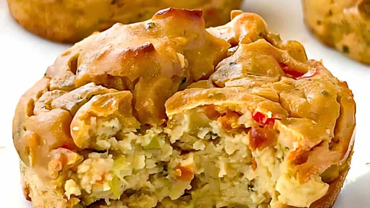 A close-up of a savory muffin with vegetables is shown with a bite taken out of it, revealing a filling with visible bits of greens, herbs, and red bell peppers. The muffin has a golden-brown crust and appears moist and flavorful.
