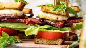 A close-up of an appetizing sandwich stack featuring layers of thick, crispy bacon, fresh lettuce, juicy tomato slices, creamy cheese, and crunchy bread garnished with fresh basil leaves on top, all secured with a skewer. The vibrant ingredients are displayed on a wooden board.