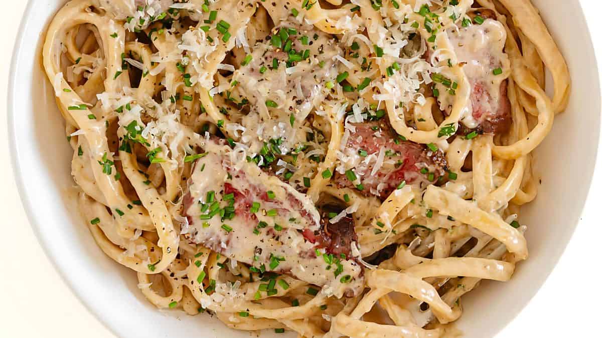 A close-up image of a creamy fettuccine Alfredo pasta dish topped with slices of grilled meat, finely chopped chives, and grated Parmesan cheese, all sprinkled with black pepper, served in a white bowl.