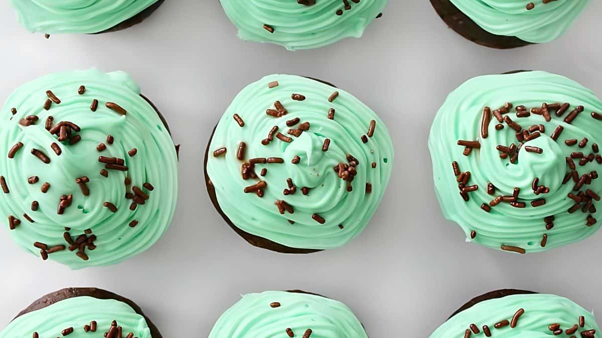 A top-down view of several cupcakes with light green frosting and chocolate sprinkles, arranged neatly on a white surface. The cupcakes are uniformly decorated, showcasing a smooth and even swirl of frosting topped with scattered sprinkles.