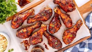 A wooden tray holds eight glazed barbecue chicken drumsticks on a sheet of parchment paper. Fresh herbs garnish the drumsticks, and a small bowl of sauce sits nearby. Fresh parsley and a bowl of coleslaw are partially visible on the sides.