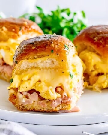 Three breakfast sliders on a white plate, each filled with scrambled eggs, ham, and melty cheese, nestled between glossy buns. Perfect for make-ahead breakfasts, they're accompanied by fresh greens and a bowl of eggs nearby, with a striped napkin and cutlery beside the plate.