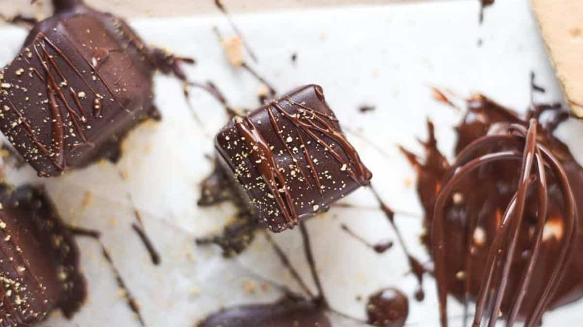 Close-up of chocolate-covered marshmallows drizzled with more chocolate and sprinkled with graham cracker crumbs, placed on parchment paper. A whisk with melted chocolate is visible on the right side, indicating dessert preparation.
