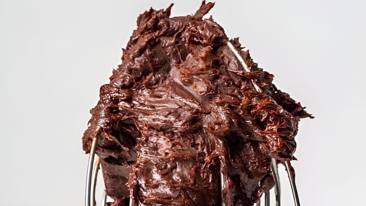 Close-up of a steel whisk covered in rich, creamy chocolate batter against a plain white background. The thick, decadent mixture clings to the wires of the whisk, highlighting its smooth texture and glossy appearance.