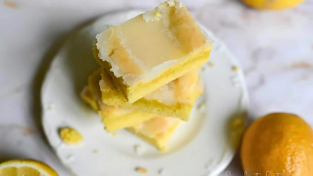 A stack of lemon bars on a white plate. The bars have a golden-yellow bottom layer and a glossy lemon glaze on top. A whole lemon is placed on the table next to the plate, and the background is blurred with a marble texture.