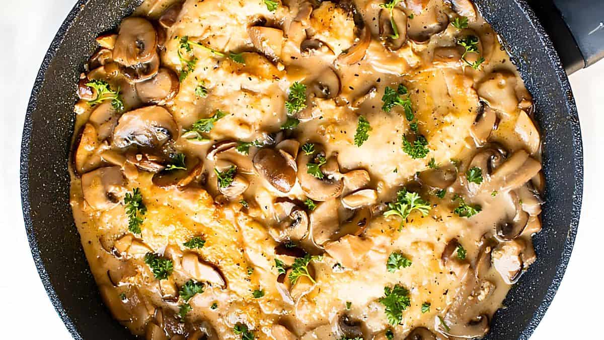 A pan filled with chicken breasts smothered in a creamy mushroom sauce garnished with fresh parsley. The dish is cooked to a golden-brown color and the mushrooms are visibly cut into slices, all covered in a rich sauce that appears well-seasoned.