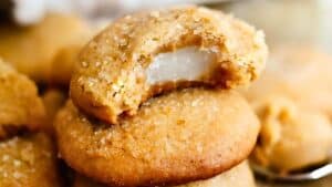 Close-up of a stack of golden-brown cookies with a sugary sheen. The top cookie has a bite taken out, revealing a soft, white filling inside. The texture appears crumbly and tender, suggesting a sweet and rich flavor.