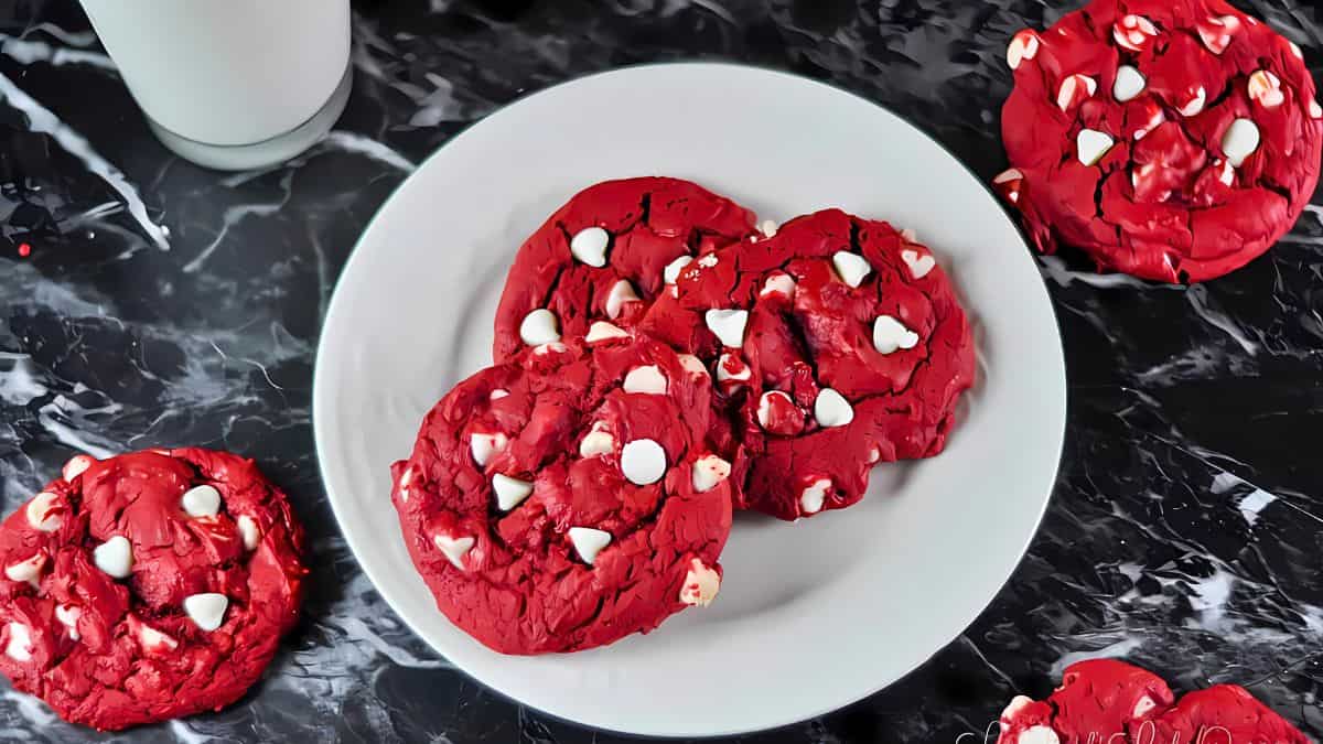 A white plate holds a stack of red velvet cookies with white chocolate chips on a black marble countertop. A glass of milk is partially visible in the background, and additional cookies are scattered around the plate.