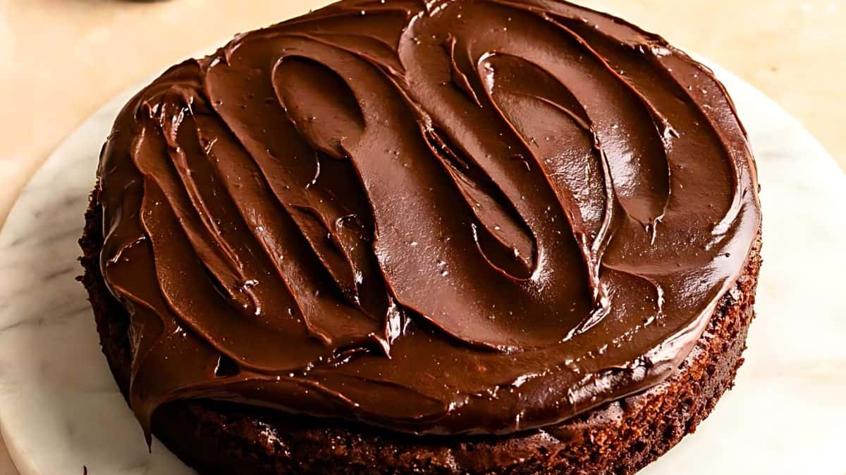 A round chocolate cake with a rich, glossy layer of chocolate frosting swirled on top. The cake sits on a marble surface, highlighting its decadent and moist appearance. The frosting is spread in artistic, wavy patterns.