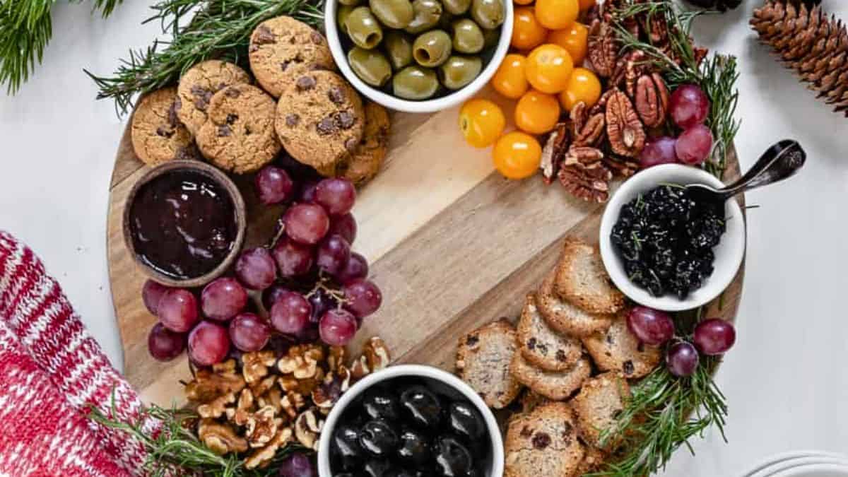 A round wooden platter featuring an assortment of snacks including green and black olives, cherry tomatoes, pecans, walnuts, grapes, chocolate chip cookies, crackers, and bowls of jam or jelly. The platter is garnished with sprigs of fresh rosemary.