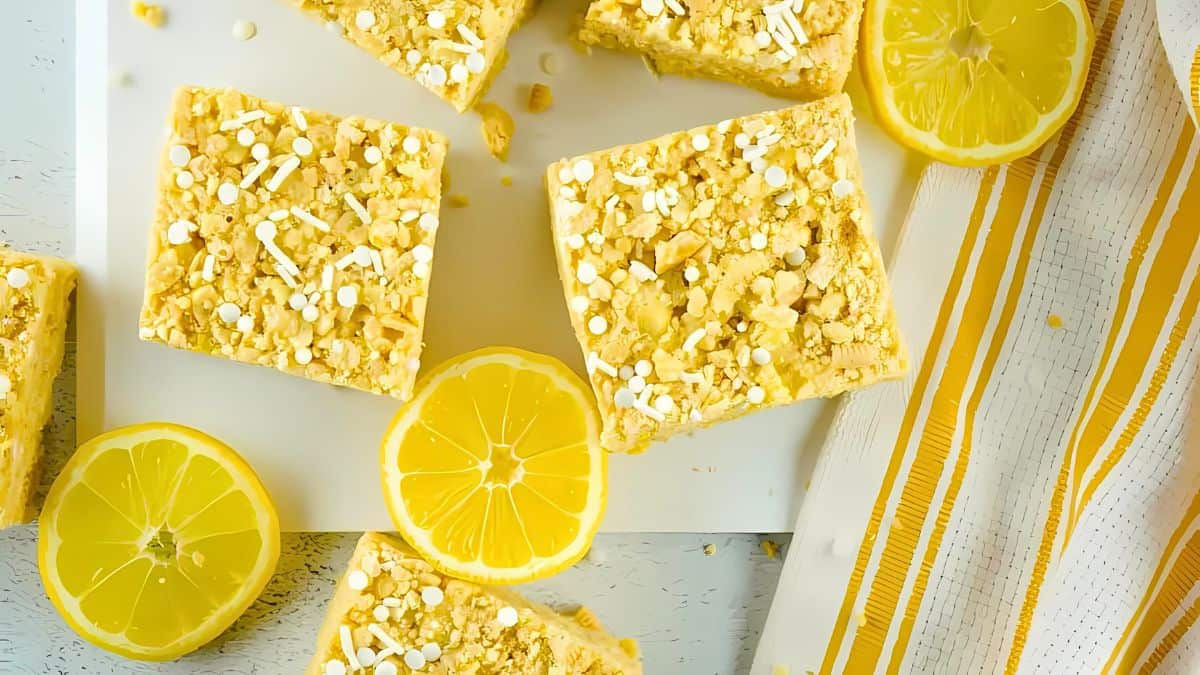 Six lemon-flavored dessert bars topped with white sprinkles and crumbs are arranged on a white surface. Two halved lemons and a yellow and white striped napkin are placed near the bars, adding a fresh, citrusy aesthetic to the scene.