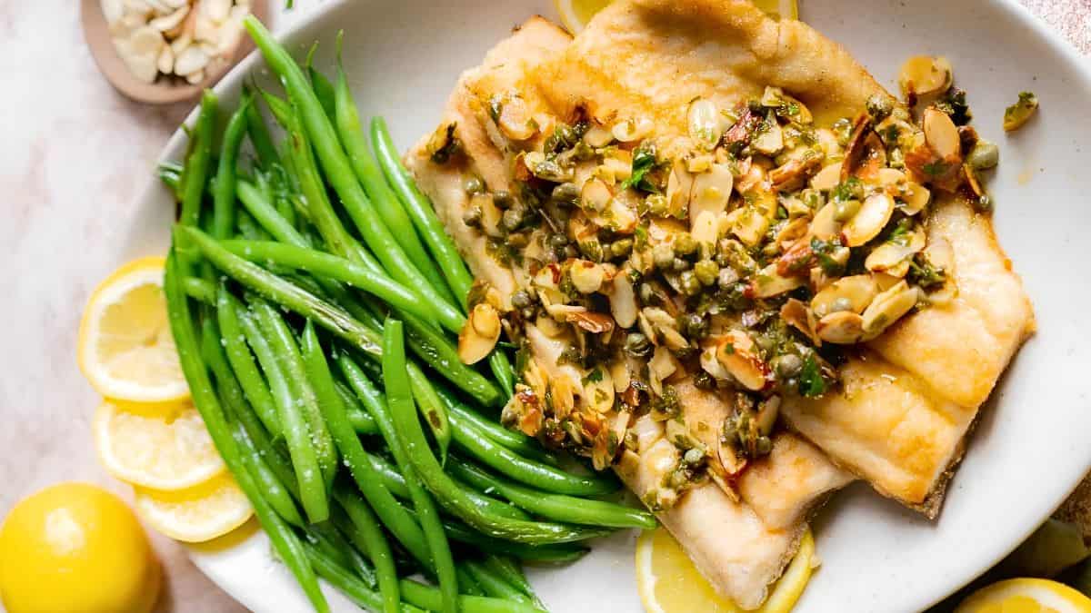 A plate of food featuring a fillet of fish topped with a mixture of sliced almonds and herbs, accompanied by a serving of green beans. Lemon slices are arranged on the side of the plate. The dish is garnished with additional almonds and herbs.