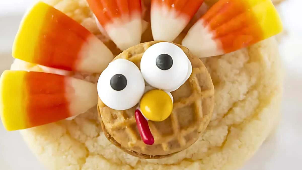 A cookie topped with a small waffle, candy eyes, and a red candy piece for a beak, resembling a turkey's face. Candy corn pieces are arranged in a fan shape, mimicking a turkey's feathers.