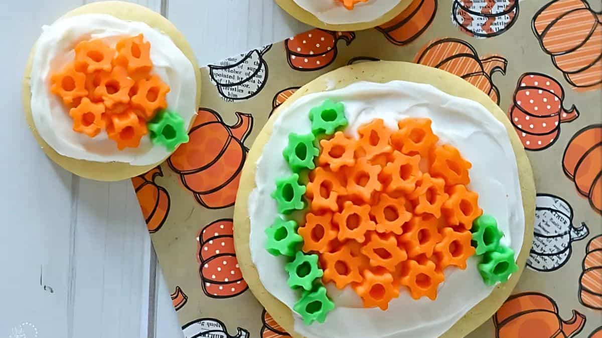 Cookies decorated with white frosting and orange flower-shaped candies, arranged to resemble pumpkins. Green candies form leaves on the sides. They are placed on pumpkin-patterned paper.