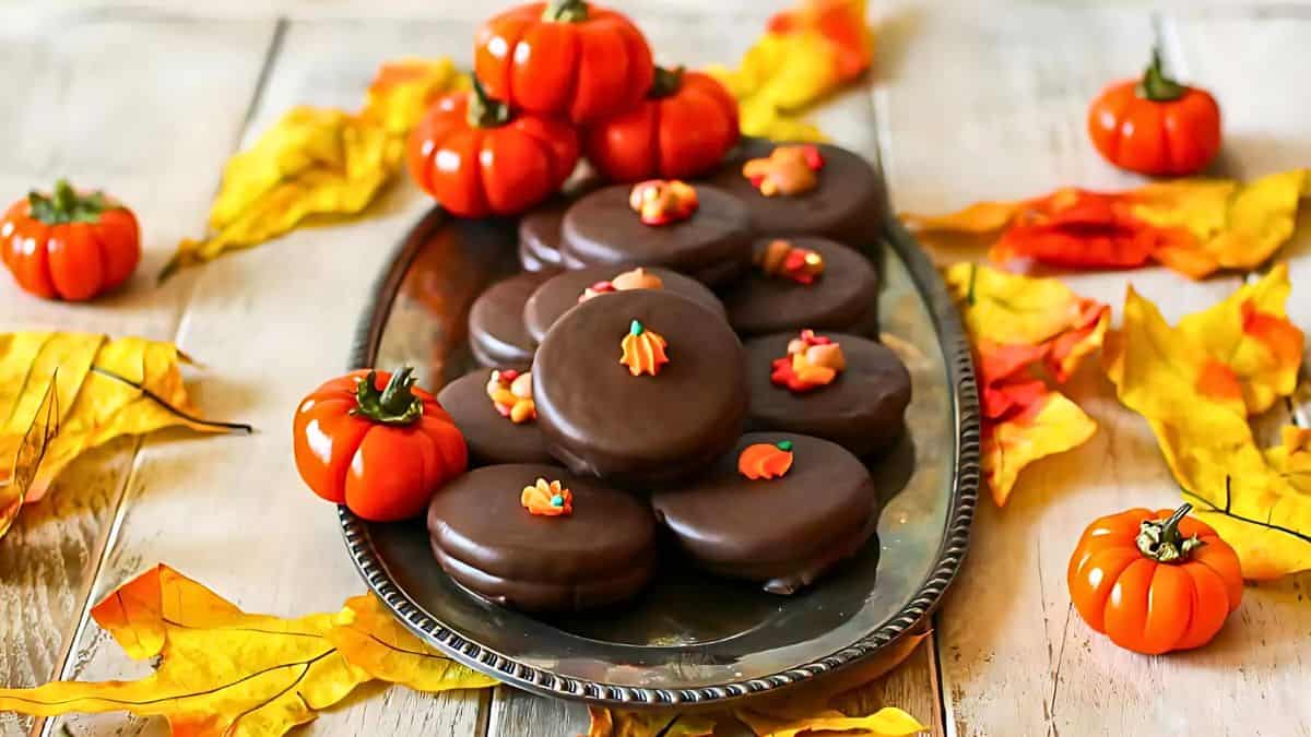 A silver platter holds chocolate-covered cookies decorated with pumpkin designs. Mini decorative pumpkins and vibrant autumn leaves surround the platter on a wooden table.