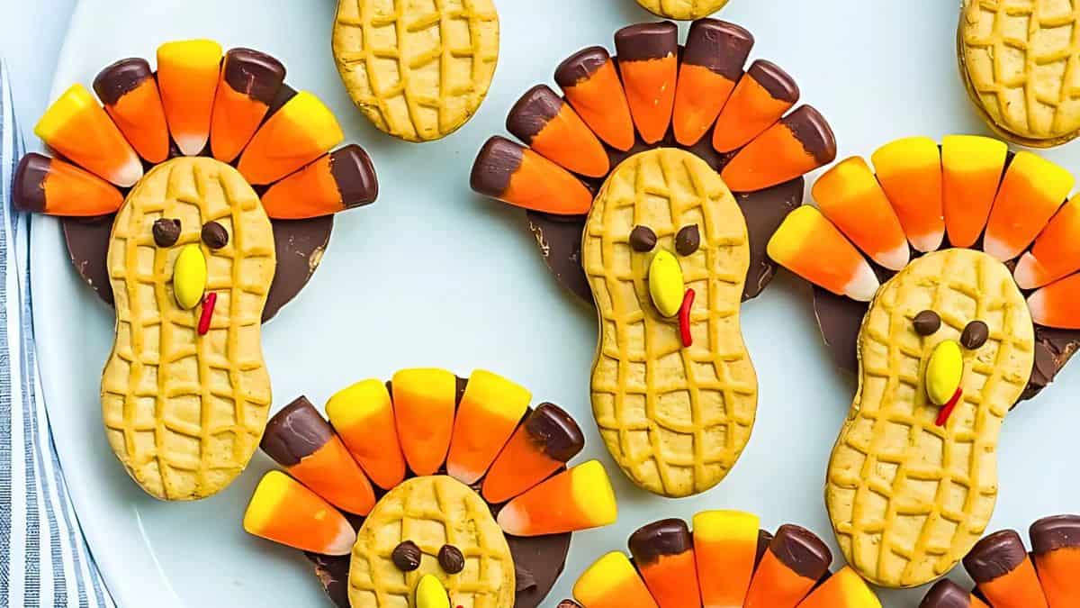 Peanut-shaped cookies decorated to look like turkeys with candy corns for feathers and chocolate chips for eyes. The cookies are arranged on a white plate.