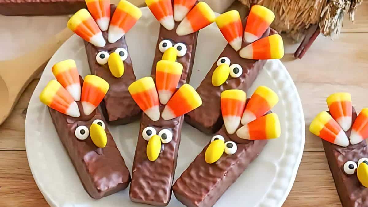 A plate of chocolate-covered treats decorated to look like turkeys with candy corn feathers and eyes made from icing, set on a wooden table.